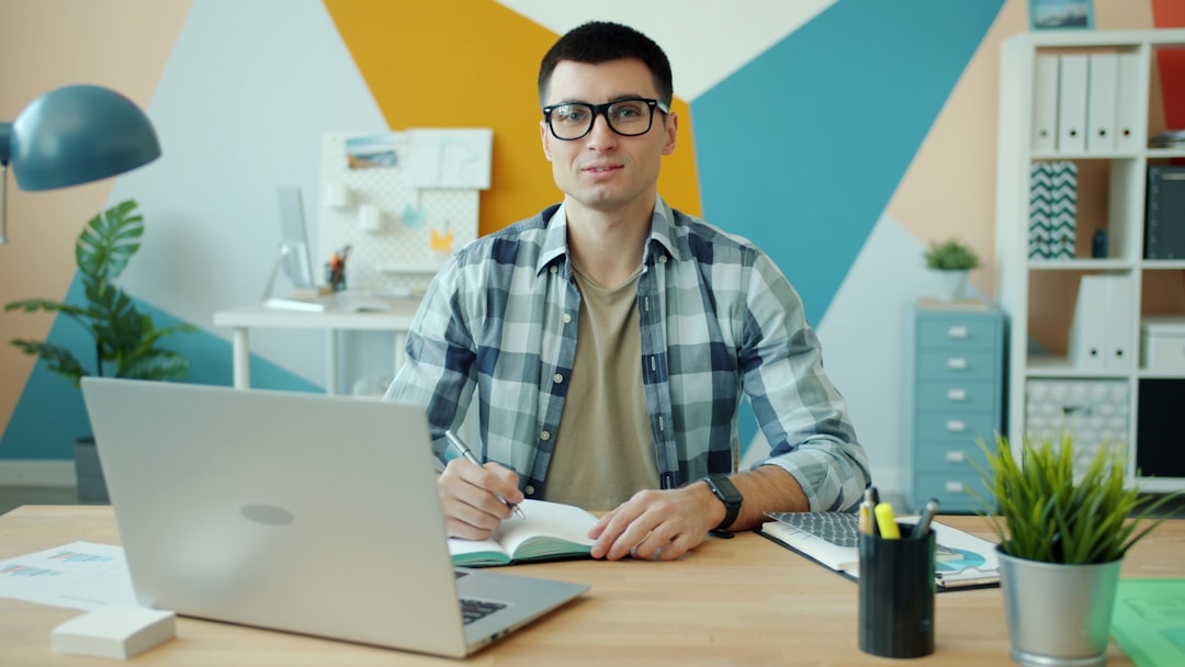 Portrait of handsome young man in glasses looking at camera sitting at desk holding pen and notebook, laptop computer is visi