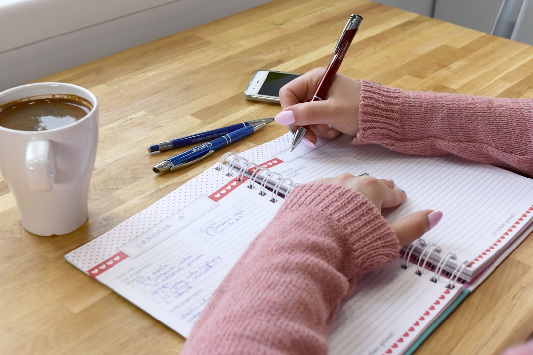 A woman in a cozy pink knitted sweater writing a recipe in a notebook on a wooden table, with a cup of coffee and pens nearby