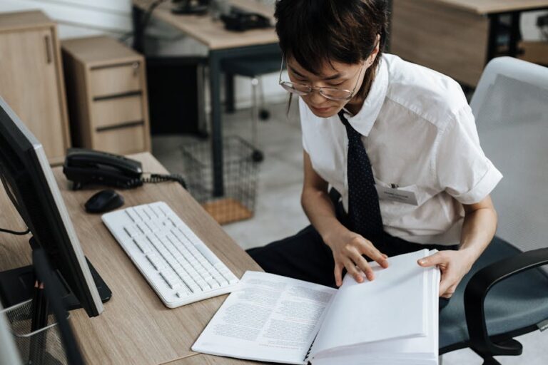 Professional in office attire reviewing documents at a desk with computer and book.
