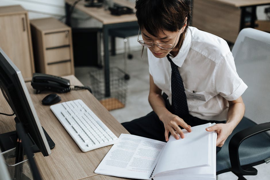 Professional in office attire reviewing documents at a desk with computer and book.