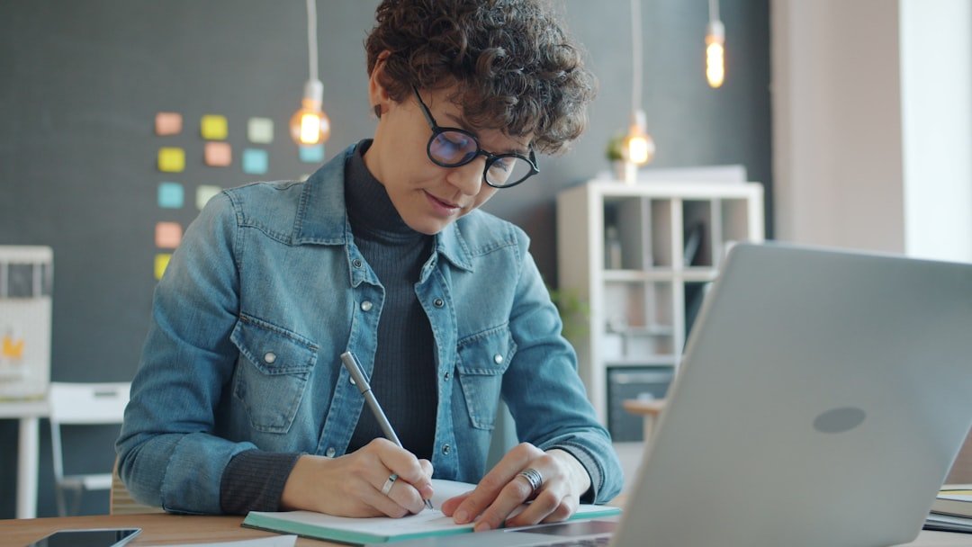 Attractive girl is using laptop and taking notes in office working alone in creative workplace focused on business activity. 