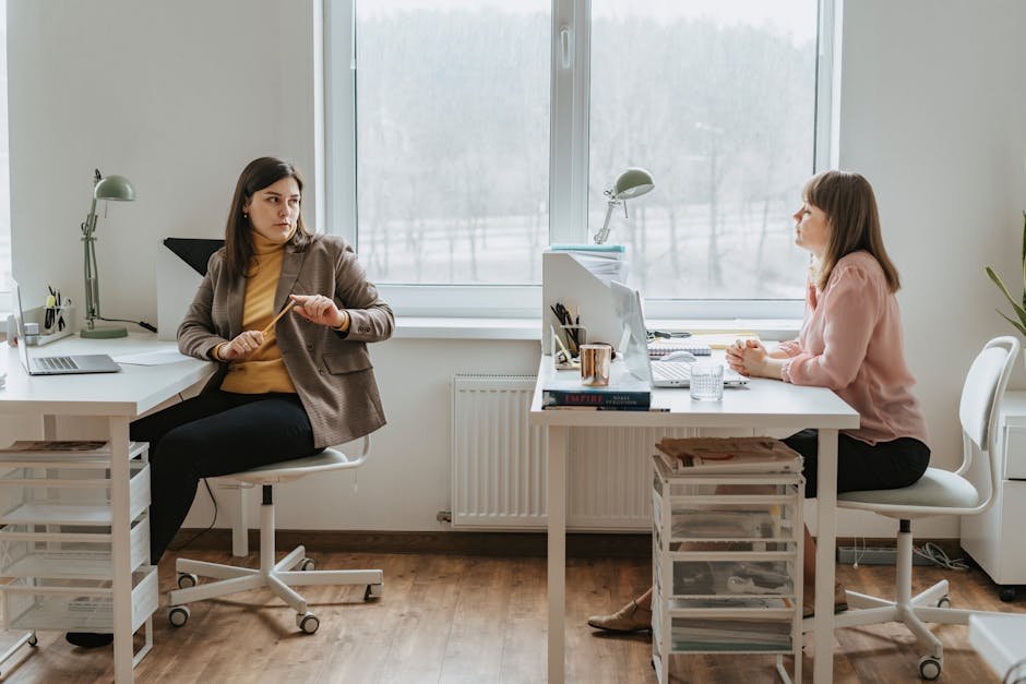 Two women having a conversation in a bright and modern workspace with desks and laptops.
