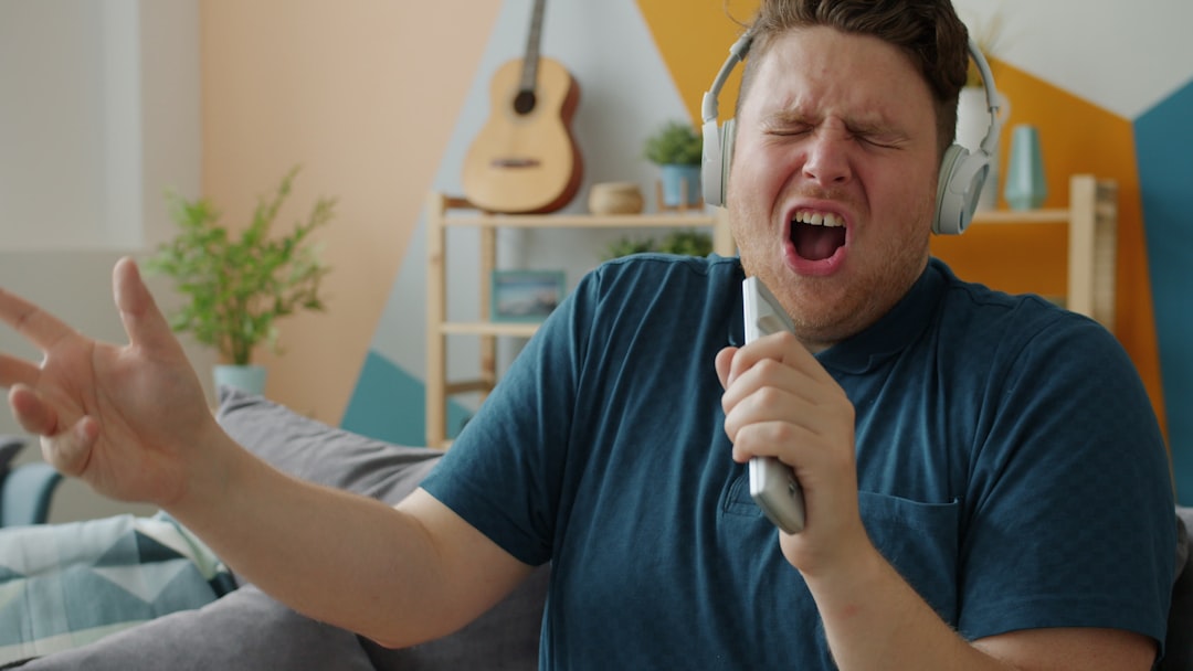 Emotional young man is singing in remote control wearing wireless headphones enjoying song indoors at home. Modern devices an