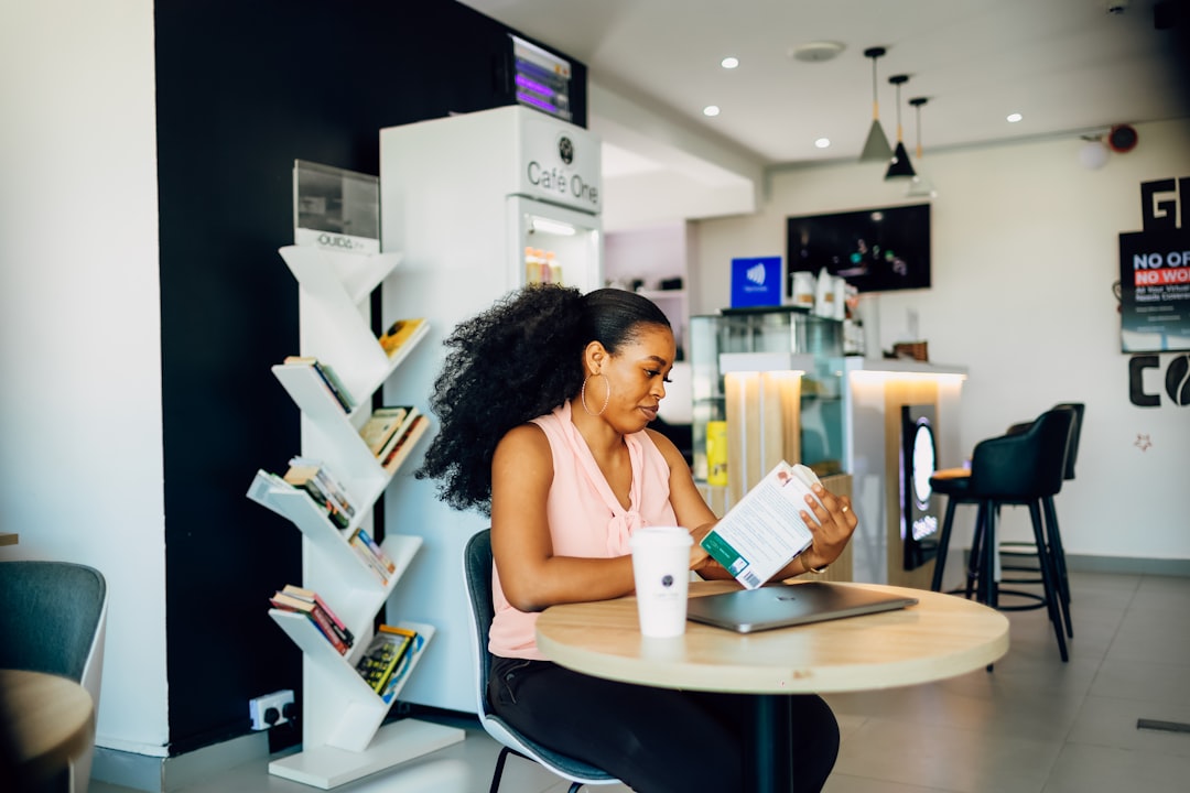 A woman sits at a table in a cafe, reading a book. She has a cup of coffee, and a laptop open on the table in front of her. F