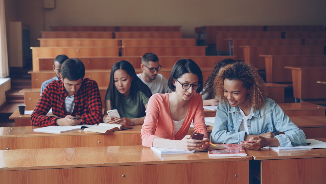 Young people are using smartphones and talking sitting at desks in lecture hall at university. Spacious light room with woode