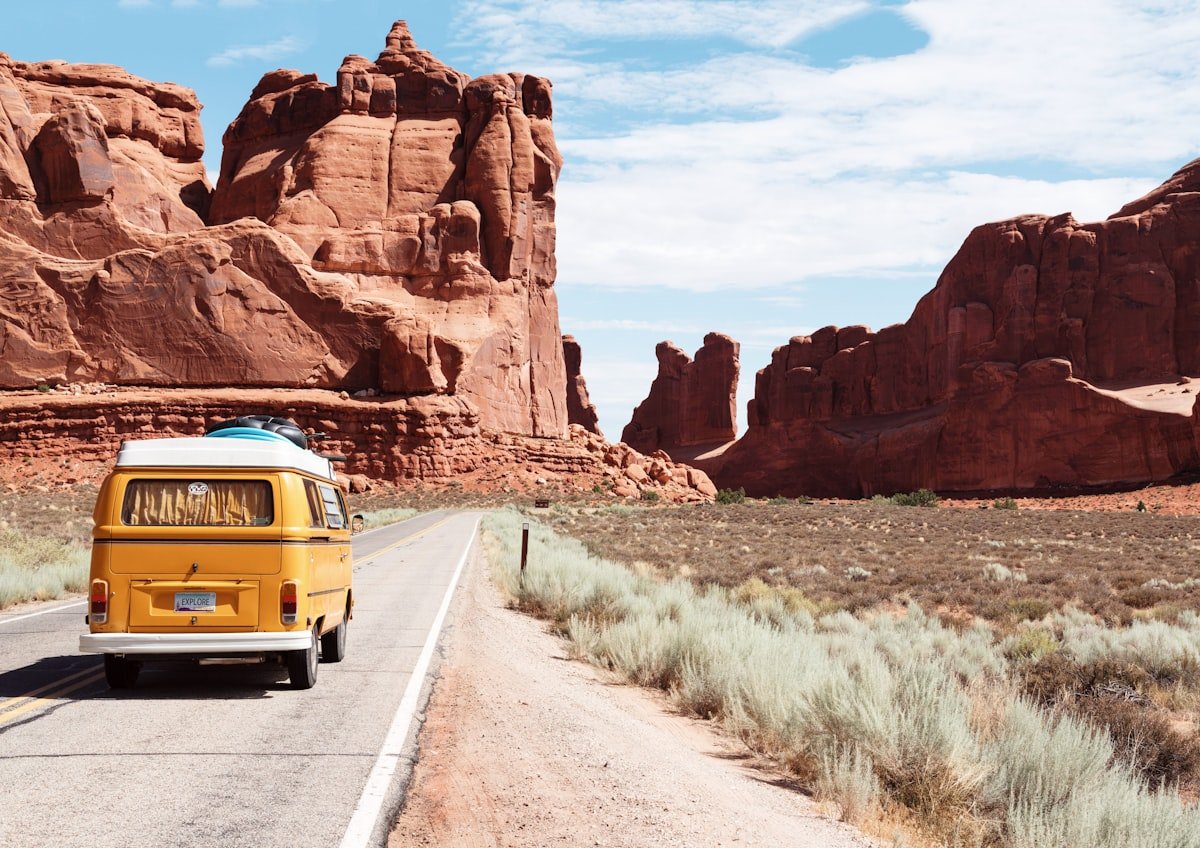 Yellow camper van driving down an open desert road past red rock formations, ready to hit the road