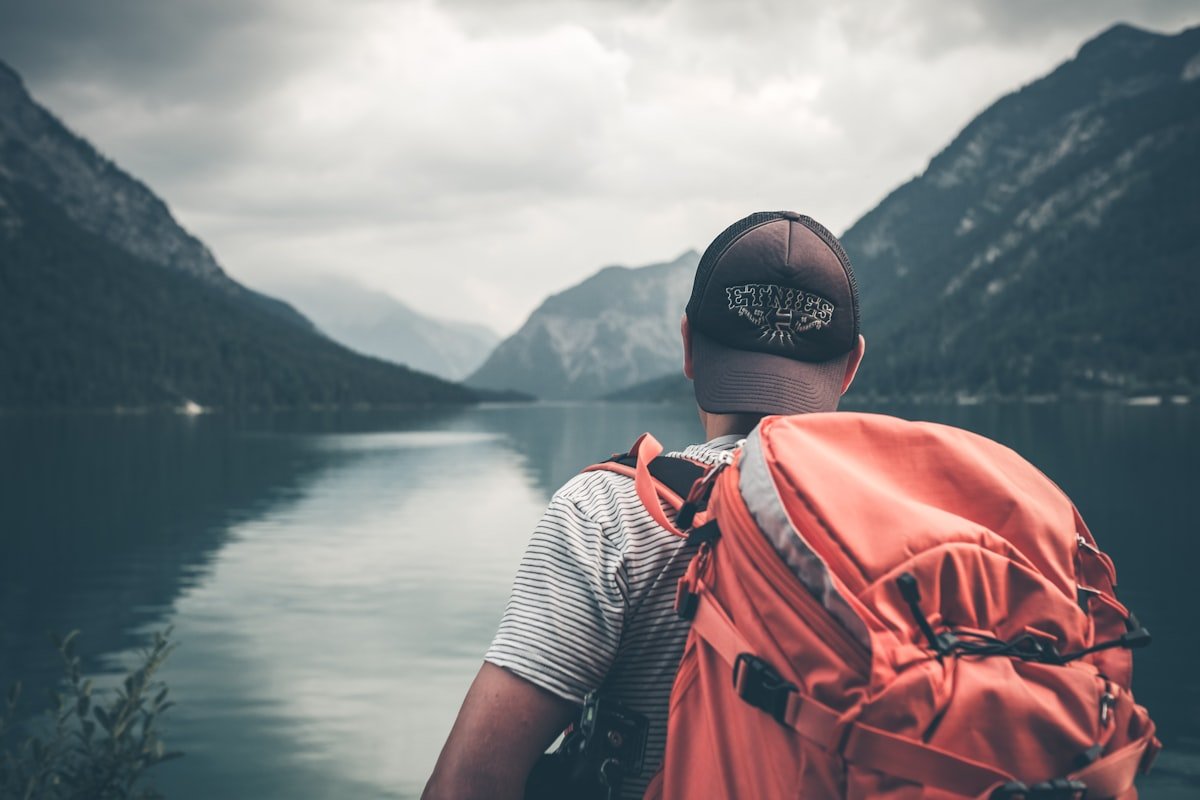 Backpacker with an orange rucksack gazing out over a misty mountain lake, itching to explore